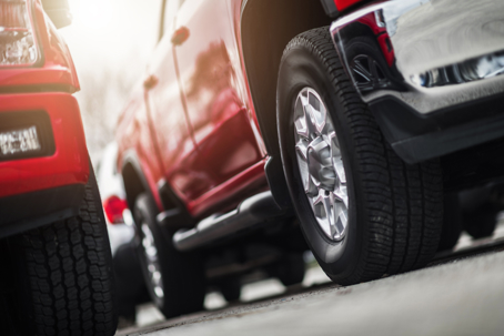 A low-angle shot captures red and silver pickup trucks parked outdoors. Sunlight illuminates the vehicles, with focus on their tires and lower bodies.